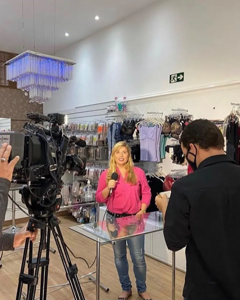 Rafaela Alcantara de blusa rosa, com microfone, dando entrevista para uma equipe de TV dentro de uma loja de roupas